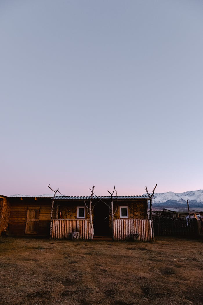 A rustic cabin in a rural setting with mountains in the background under a twilight sky.